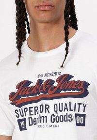 White cotton t-shirt featuring a bold printed logo in navy and red, stating "Jack & Jones" and "Superior Quality Denim Goods."