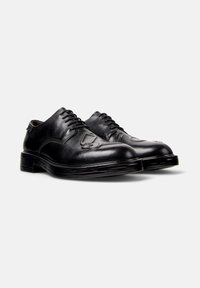 Un paio di scarpe eleganti in pelle nera con lacci, con design impresso sulla punta, suola spessa e tacco basso su uno sfondo bianco semplice.