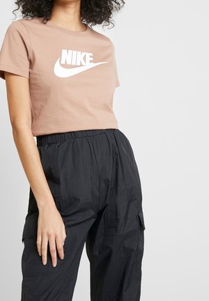 Vrouw draagt een lichtbruine Nike-t-shirt met wit logo en losse zwarte cargobroek tegen een effen lichte achtergrond.