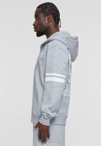 Grå hoodie med frontlomme, snøre i hætten og hvide striber på ærmerne. Fremstillet af blødt, tekstureret stof.