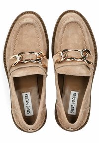 Mocasines de ante en beige con un detalle de cadena dorada en la parte delantera, con un borde cosido y una suela texturizada. Diseño plano, sin tacón.