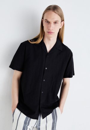 Juodas trumpas rankovės mygtukų marškinėlis, pagamintas iš lengvos medžiagos, su klasikiniu apykaklės kirpimu ir subtilia tekstūra, derinamas su dryžuotomis kelnėmis.