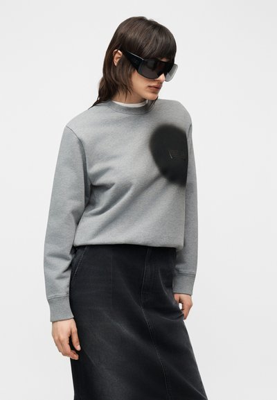 Άτομο που φοράει μαύρα υπερμεγέθη γυαλιά ηλίου, γκρι φούτερ με μαύρο λογότυπο και μαύρη φούστα ψηλής μέσης, σε λευκό φόντο.