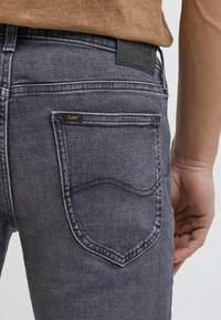 Jeans de mezclilla gris con un diseño de bolsillo trasero cosido y una etiqueta de cuero con el nombre de la marca. Primer plano de una mano en la cintura.