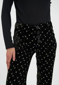Pantalon taille haute en velours noir avec des motifs floraux dorés, fermetures à cinq boutons à l'avant et deux poches latérales. Texture douce et coupe ajustée.