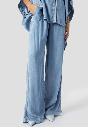 Person som har på seg løse, lyseblå denim-bukser og matchende oversized skjorte med knapper, med hvite sko med spiss tå, stående på grå bakgrunn.