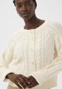 Kreminės spalvos kabelinio rašto kardiganas su tekstūriniais raštais, raštuotomis rankovėmis ir penkiais mygtukais priekyje. Minkšta audinio medžiaga.