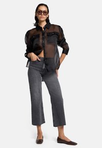 Schwarze, durchsichtige Bluse mit zwei Brusttaschen, kombiniert mit dunkelgrauen, cropped Jeans und braunen flachen Schuhen. Das Modell trägt übergroße Sonnenbrillen.