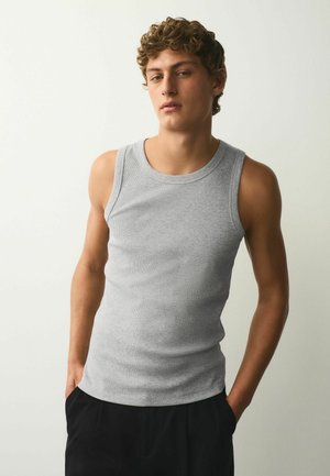 Młody mężczyzna z kręconymi włosami, ubrany w szary bezrękawnik i czarne spodnie, stoi z rękami w kieszeniach na jednolitym tle.