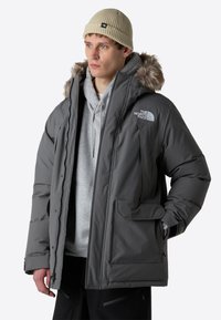 Grå isolerad jacka med huva i fuskpäls, dragkedja och knäppning, flera fickor, bärs över en grå hoodie med en logotyp.