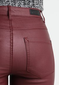 Pantalones de imitación piel en color burdeos con un diseño ajustado, detalles cosidos y un pequeño bolsillo trasero como acento. Presentan una textura lisa y brillante.