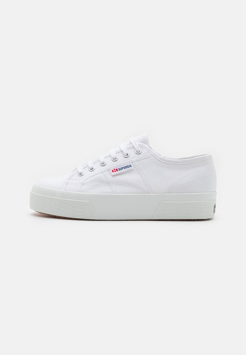Hvite lerrets sneakers med teksturert gummisåle, snøring og et rødt og blått logo-patch på siden. Enkel, lavprofil form.