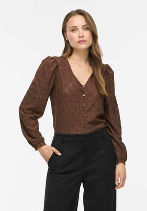 Femme aux longs cheveux portant un chemisier marron texturé boutonné avec des manches bouffantes et un pantalon noir sur mesure, debout avec une main dans la poche.