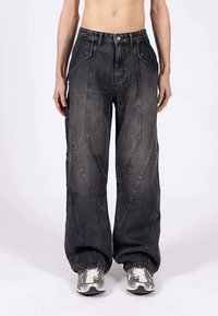 Άτομο που φοράει φαρδιά μαύρα τζιν παντελόνια με καμπύλες ραφές και ασημί-γκρίζα αθλητικά παπούτσια, που στέκεται μπροστά σε ένα απλό φόντο.