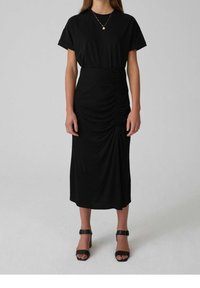 Abito midi nero con scollatura tonda, maniche corte e dettagli arricciati sui lati; abbinato a tacchi neri con cinturini.