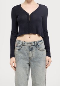 Granatowy prążkowany sweter na zamek, skrócony z długimi rękawami, z głębokim dekoltem w kształcie litery V i teksturowanym materiałem, zestawiony z wyblakłymi jasnoniebieskimi dżinsami.