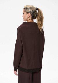 Vrouw met een blonde paardenstaart, gekleed in een oversized donkerbruine trui met lange mouwen en bijpassende broek, die naar links kijkt tegen een effen witte achtergrond.