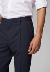 Homme portant une chemise blanche à boutons et un pantalon texturé bleu foncé, la main détendue sur le côté contre un fond uni.