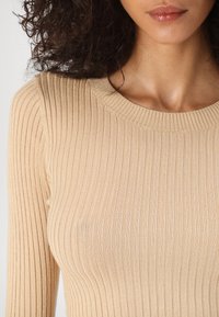 Haut en maille côtelée beige avec un col rond et des manches longues, offrant une texture douce et un éclat subtil.