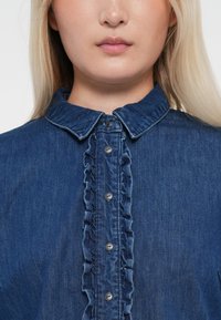 Sinine teksasärk, millel on volangiga esine, nööbikeeramine ja klassikaline krae. Kude on pehme ja tekstuuriga viimistlusega.