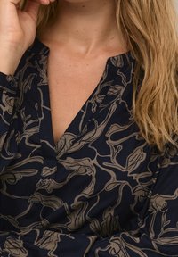 Blouse en tissu bleu marine avec motif floral beige clair, dotée d'un décolleté en V et d'un détail à boutons sur la manche. Texture lisse partout.