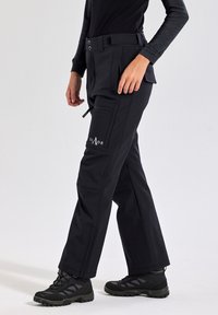 Zwarte, duurzame softshell broek met cargozakken, elastische tailleband en ritsen. Draag ze met robuuste zwarte wandelschoenen voor buitenactiviteiten.