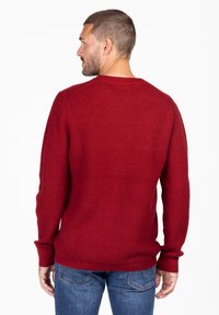Rode gebreide trui met een gestructureerd patroon, ronde hals en lange mouwen. Gecombineerd met blauwe jeans, met de achterzijde zichtbaar.
