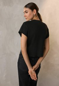 Černý zkrácený top s krátkými rukávy, vyrobený z hladkého materiálu. Model nosí černé džíny. Jednoduchý design bez viditelných vzorů nebo ozdob.