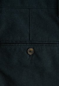 Mørk denimstof med en fin tekstur, der har en flad lomme og en rund, fire-huls knap. Syninger tilføjer subtile designaccenter.