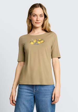 Blondynka w beżowej koszulce z krótkim rękawem z grafiką cytryny i szklanki, w połączeniu z niebieskimi dżinsami, stoi na jasnym, jednolitym tle.