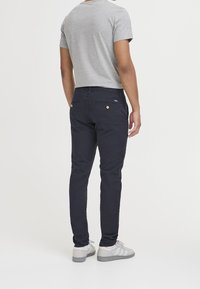 Pantaloni slim fit blu navy con tasche posteriori e bottoni color marrone chiaro, abbinati a una camicia grigia a maniche corte e sneaker grigie.