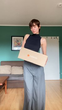 Kartónová krabička Zalando držená pred osobou v bezrukávovom čiernom topu a svetlošedých širokých nohaviciach, s zelenou stenou za ňou.