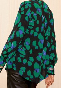 Blusa negra con manchas abstractas verdes y azules, de mangas largas y detalles en los puños. El material parece ligero y texturizado.