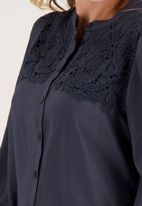 Marineblauwe blouse met knopen en kantdetails op de schouderpartij. De stof heeft een gladde textuur, met een kraag en lange mouwen.