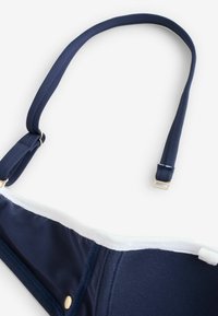 Tmavomodrý ramienkový popruh podprsenky so zlatým nastaviteľným kovovým dielom a bielym lemovaním na bielom pozadí.