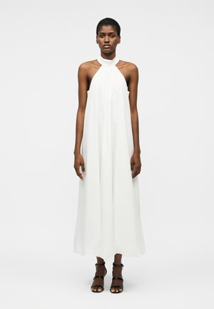Femme portant une robe maxi blanche sans manches à col licou et des sandales noires à talons hauts à lanières, debout devant un fond uni.