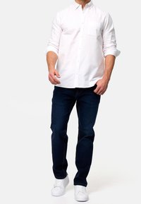 Witte overhemd met knopen en een borstzak, gecombineerd met donkerblauwe jeans en witte sneakers, voorzien van een getailleerd ontwerp en opgerolde mouwen.