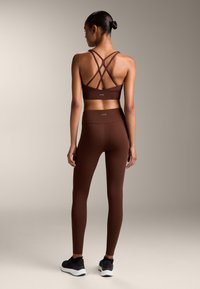 Hnedý športový podprsenka s kríženými ramienkami, spárovaná s hnedými legínami s vysokým pásom. Model má na sebe čierne športové topánky. Hladká, matná textúra.