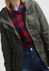 Olivově zelená bunda s kapucí a zipem vpředu, s nastavitelným pasem a stříbrnými detaily, vrstvená přes červenou a modrou kostkovanou košili.