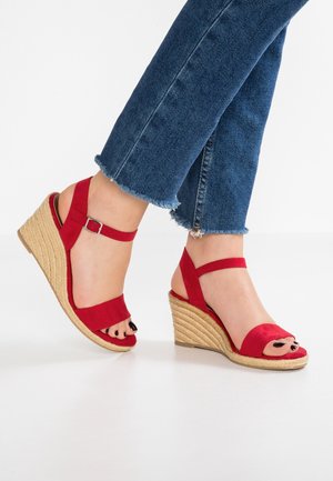 Pieds portant des sandales compensées rouges avec des brides à la cheville et un jean bleu à ourlet effiloché sur fond blanc.