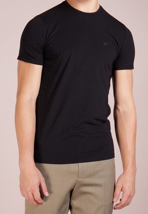 Man draagt een nauwsluitend zwart T-shirt met korte mouwen en ronde hals, gecombineerd met beige broek, staand tegen een effen achtergrond.