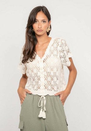 Femme aux longs cheveux foncés portant un haut blanc en crochet à boutons et un pantalon vert olive avec des cordons à pampilles, debout devant un fond uni.