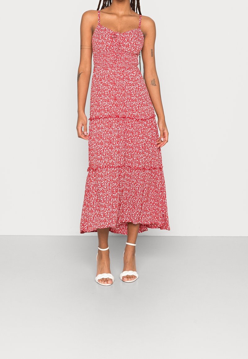 Femme portant une robe midi rouge sans manches avec un petit imprimé floral blanc et des sandales blanches à talons avec bride à la cheville, debout devant un fond uni.