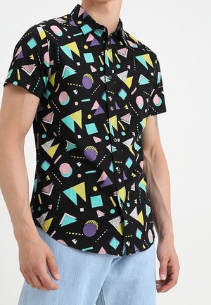 Mann trägt ein schwarzes Kurzarmhemd mit bunten geometrischen Formen und hellblauen Hosen, steht vor einem weißen Hintergrund.