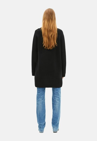 Schwarzer, gestrickter Oversize-Pullover mit langen Ärmeln, gerippten Bündchen und Saum, kombiniert mit hellblauen, figurbetonten Jeans. Lange, wellige Haare sind sichtbar.