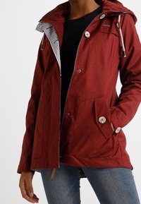 Giacca con cappuccio rosso realizzata in tessuto testurizzato. Presenta una foderatura a righe contrastanti, due ampie tasche e bottoni in legno per la chiusura.