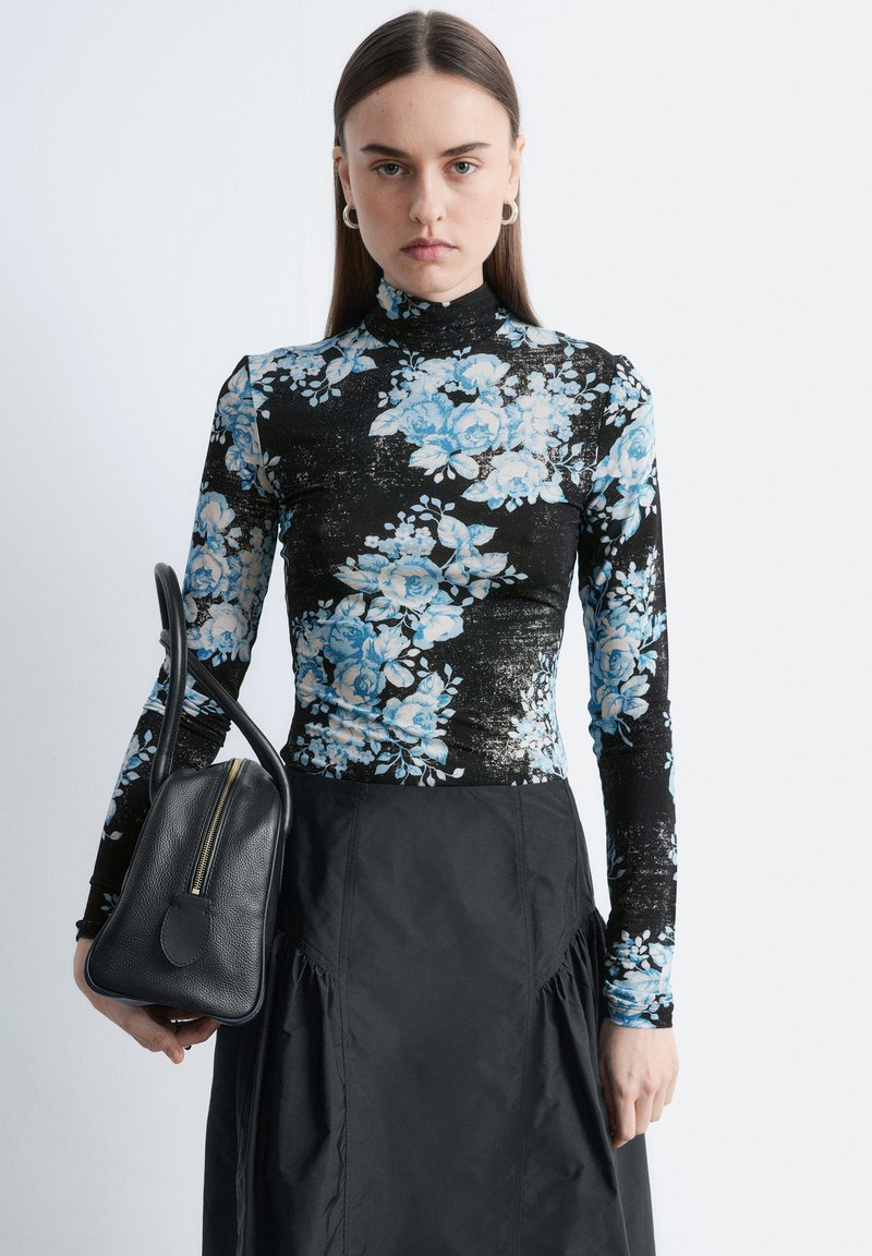 Femme portant un col roulé noir à imprimé floral bleu et une jupe noire, tenant un sac à main en cuir noir avec une fermeture éclair dorée.