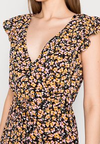 Femme portant une robe noire avec un motif floral rose, orange et blanc, décolleté en V profond, manches flottantes et une ceinture à la taille.