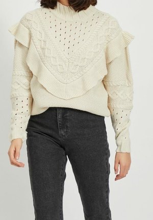 Pull crème en maille torsadée avec empiècement à volants et détails perforés sur les manches, associé à un jean noir taille haute.