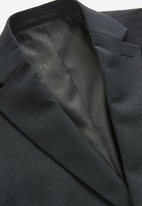 Dunkelblazer aus strukturiertem Stoff, mit einem Reverskragen und einem gemusterten schwarzen Innenfutter, das sichtbar ist.